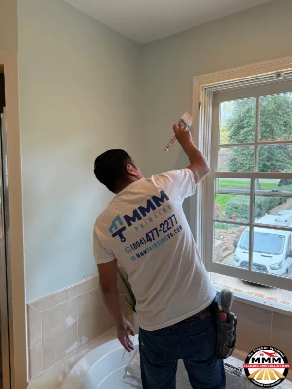 Worker applying paint to bathroom wall above tile wainscoting near window